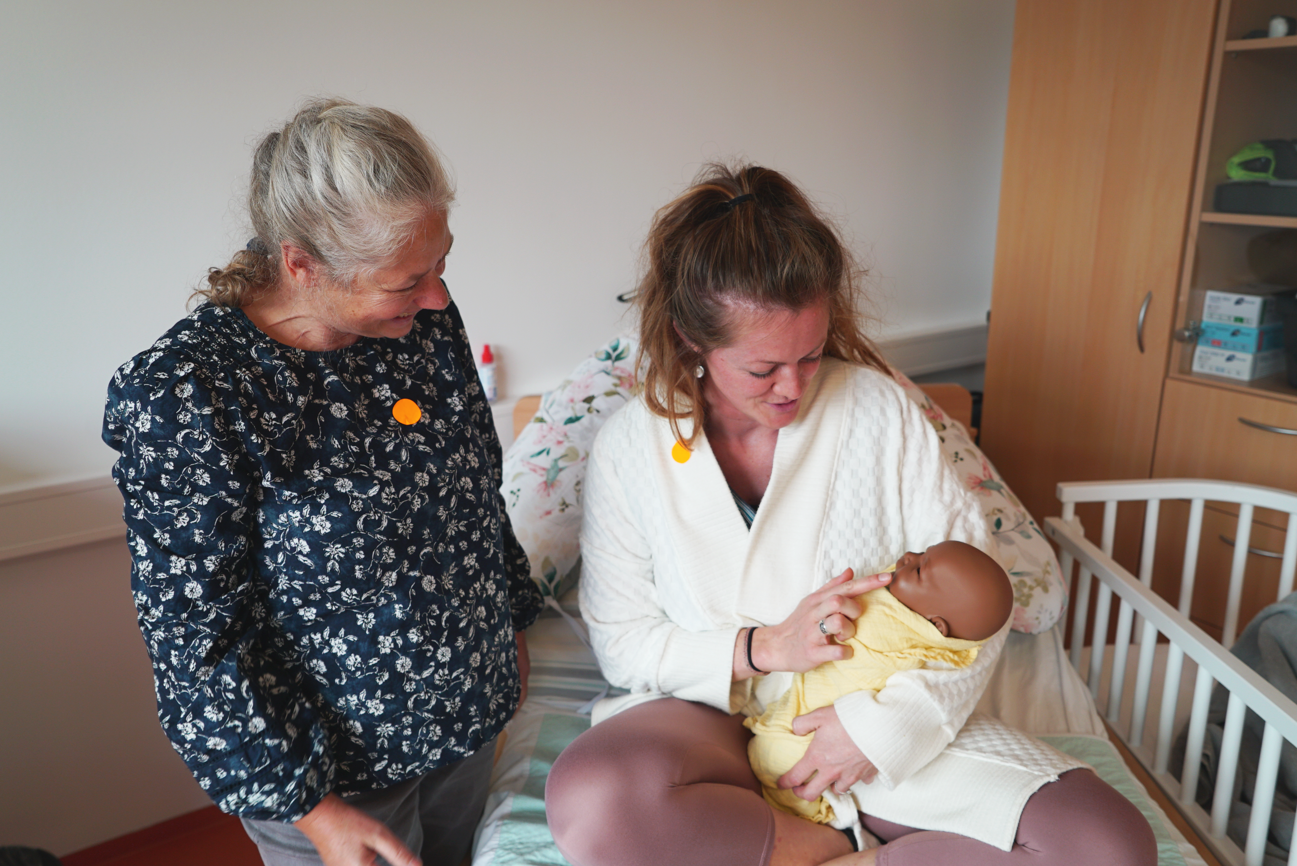 Two women, one of whom is sitting on a hospital bed holding a baby doll