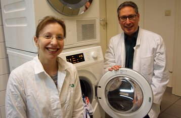 A woman and a man in lab coats stand in front of an open washing machine
