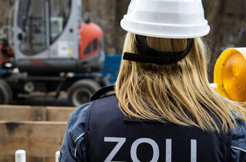 Eine Frau mit Zoll Schriftzug und weißem Helm schaut auf eine Baustelle