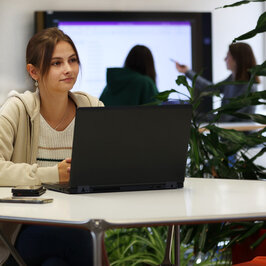 Person sitzt am Laptop, im Hintergrund zwei Personen am Smartboard