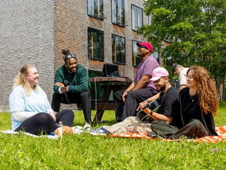 Studierende sitzen auf einer grünen Wiese vor einem Hochschulgebäude 