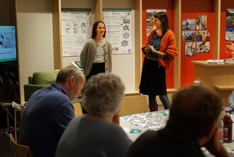 Symbolbild: Zwei Frauen stehen vor einem sitzenden Publikum