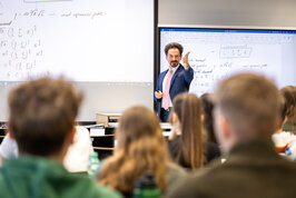 Ein Professor an einer Leinwand zeigt in die Studierenden Menge