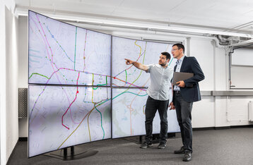 Man sieht zwei Männer die vor einer Straßennetzkarte auf TV-Bildschirmen stehen