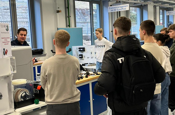 Man sieht eine Schulklasse die im Halbkreis um einen Vortragenden steht