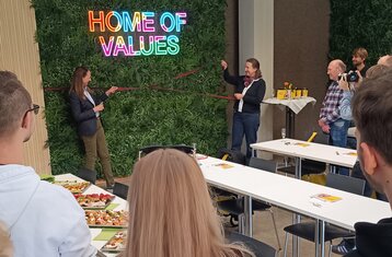 zwei Personen ziehen eine rote Schleife vor einer begrünten Wand. 