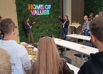 zwei Personen ziehen eine rote Schleife vor einer begrünten Wand. 