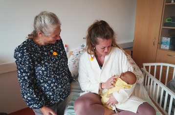 Zwei Frauen eine hält eine Babypuppe im Arm und sitzt auf einem Bett,