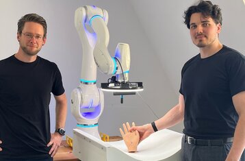 Zwei Wissenschaftler stehen vor einem Roboterarm und zeigen ein Handmodell