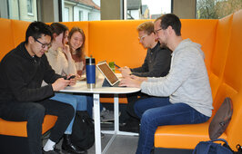 Man sieht Studierende auf einer orangenen Couch an einem Tisch sitzen