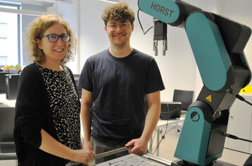Professorin und Doktorand stehen an einer Maschine mit Roboterarm