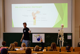 Ein Vorlesungssaal mit Kindern vorne steht ein Skelett