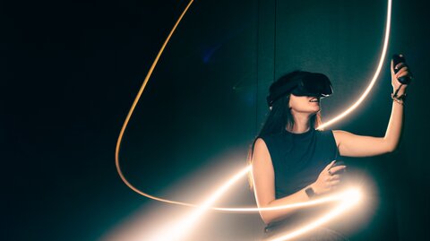 Symbolbild: Eine Frau mit VR Brille ist von Lichtbögen umgeben