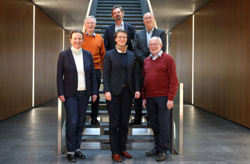 Vorne zwei Frauen ein Mann, hinten stehen 3 Männer auf einer Treppe